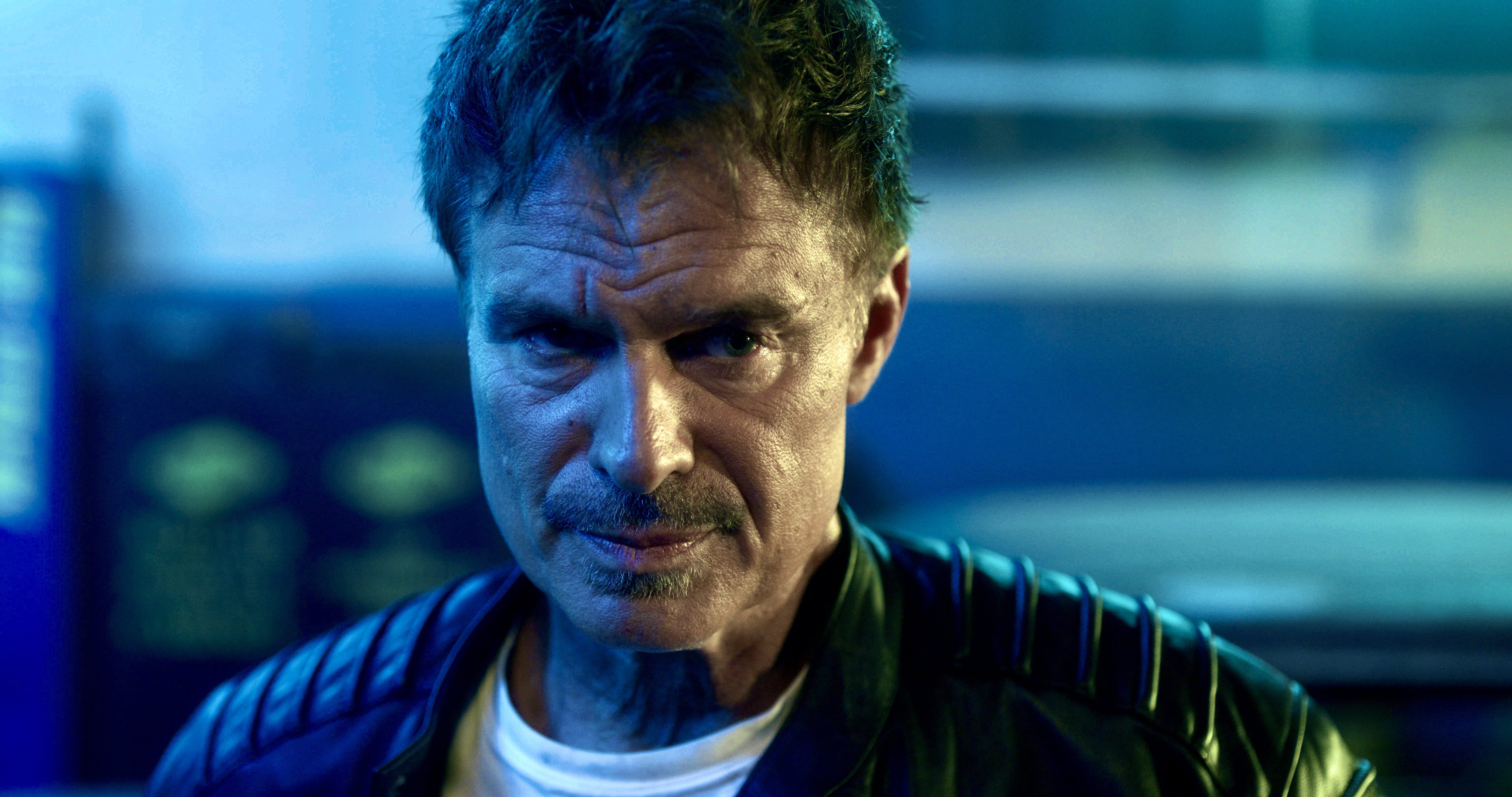 A serious-looking man with a moustache, wearing a black leather jacket, is staring intently at the camera with a blue and green lit background.