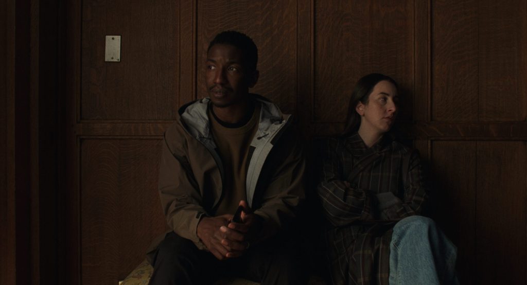 A man and a woman sit on a bench against a wooden wall, appearing distant from each other. The man wears a dark jacket and looks contemplative, while the woman, dressed in a striped coat, gazes to the side with a serious expression.