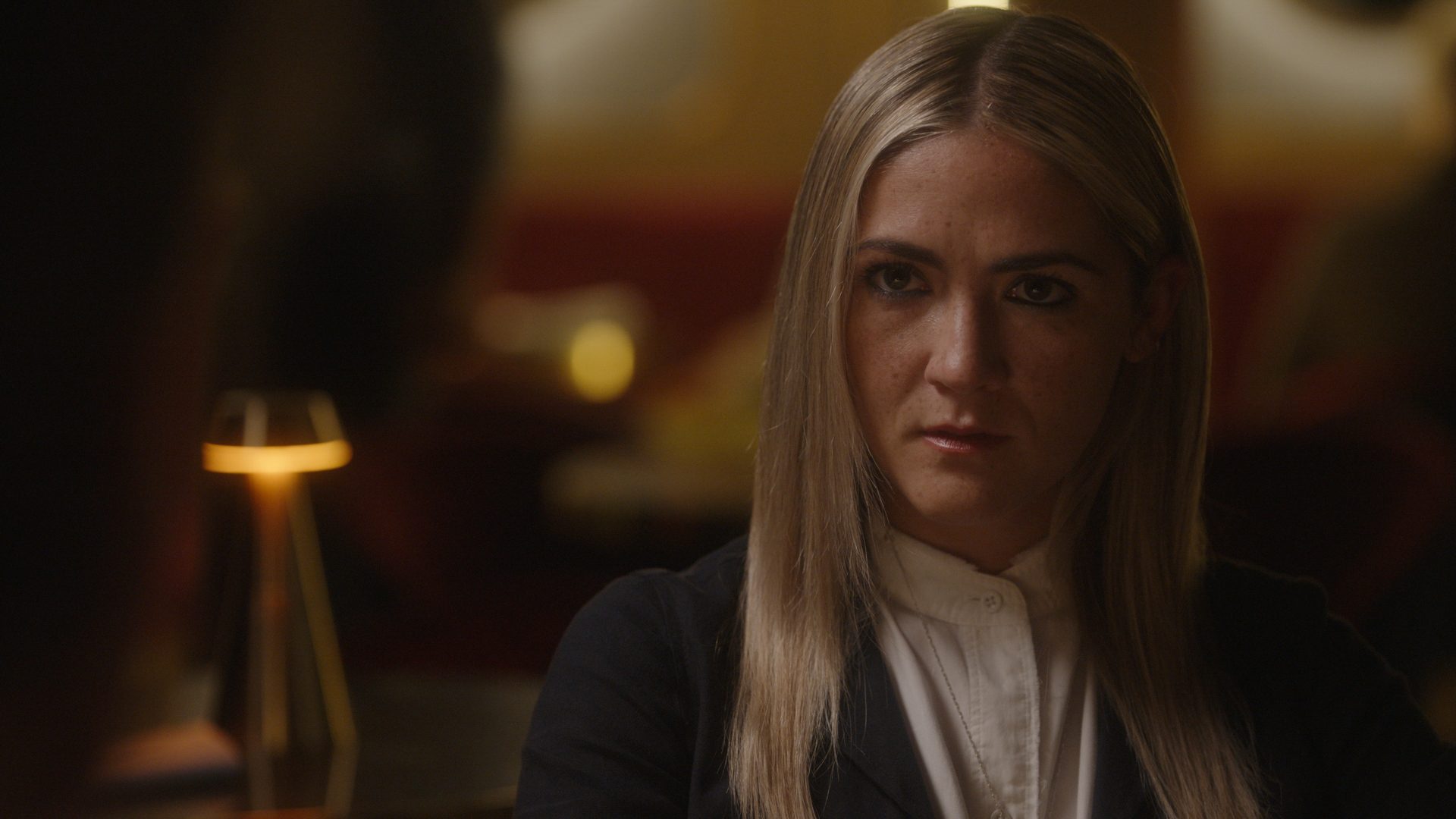 A woman with long blonde hair, wearing a black blazer and white blouse, gazes intently at someone off-camera, with a serious expression in a dimly lit setting.