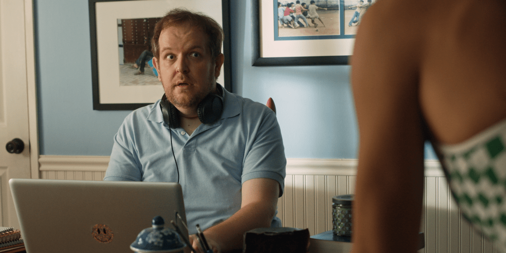 A surprised man wearing headphones sits at a desk with a laptop, looking towards an off-screen person, in a room with blue walls and framed pictures.